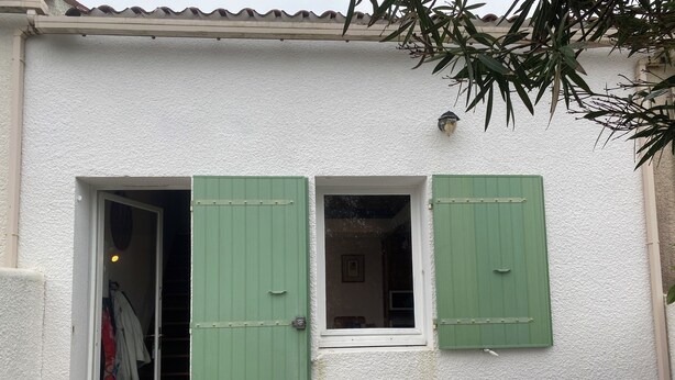 A building with a white textured exterior, two green shutters, a small window, and a door leading to a room with a bed and a wardrobe. - Vacation home located 300m from the beach (Dolus-d'Oléron)