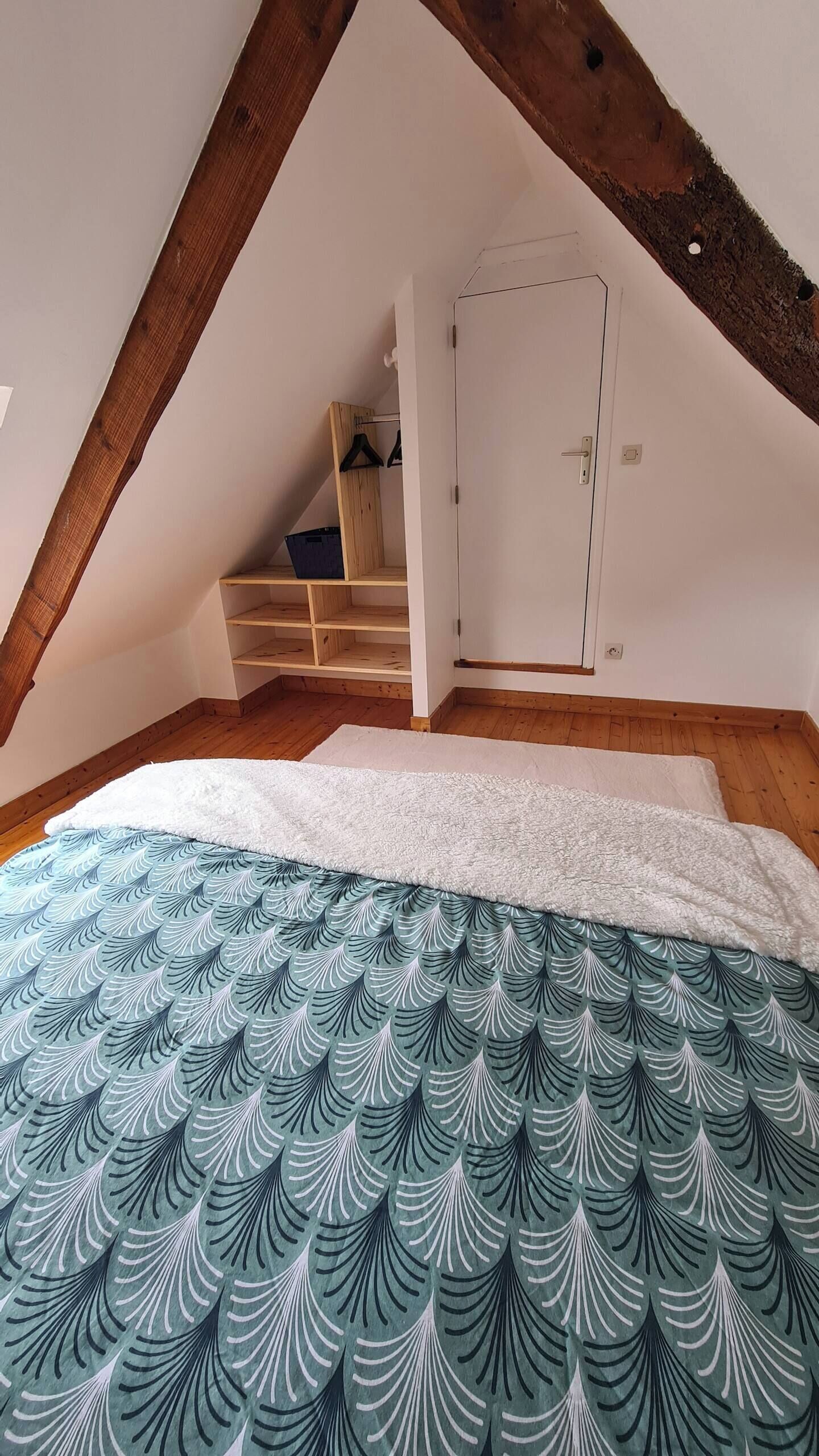 A bedroom with a sloped ceiling, wooden beams, a bed with a patterned duvet, a wardrobe, and a small shelving unit.