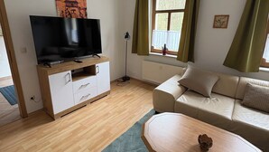 Moderni olohuone, jossa on seinälle asennettu taulutelevisio puisen lipaston päällä, beige sohva, puinen sohvapöytä ja ikkuna, jossa on vihreät verhot.