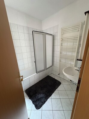 A bathroom with white tiled walls, a black rug, a white sink, and a shower with a white door.