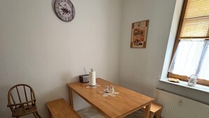 Pieni, viihtyisä huone, jossa on puinen pöytä ja penkit, patteri, ikkuna, jossa on sälekaihtimet, ja seinäkello.
