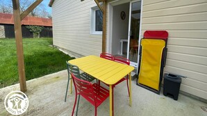 Eine überdachte Terrasse mit einem gelben Tisch und roten Stühlen, einem gelben Klappstuhl und einer schwarzen Mülltonne.