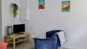 Ein modernes Wohnzimmer mit einem Flachbildfernseher, einem Holztisch, einem blauen Sessel und zwei Wänden mit Reiseplakaten.