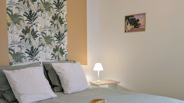 Ein ordentlich bezogenes Bett mit Kissen, eine Nachttischlampe, ein gewebter Korb und ein Tablett mit Gläsern und einer Rose in einem Hotelzimmer.