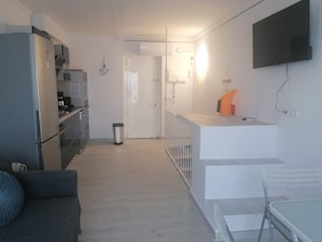 Une cuisine compacte équipée d’un réfrigérateur, d’une cuisinière et d’un évier, un petit coin repas avec une table et des chaises, et un téléviseur fixé au mur.