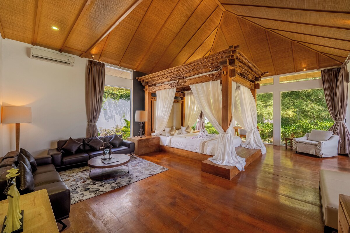 A spacious room with a wooden ceiling, a four-poster bed, a sofa, a coffee table, and a view of greenery outside.