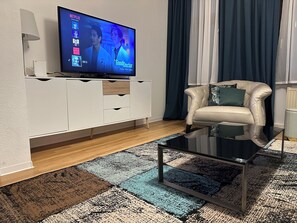 En moderne stue med et fladskærms-tv, et hvidt tv-møbel, en beige sofa med en grøn pude og en tæppe med et mønster.