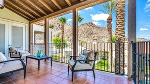 A covered outdoor seating area with chairs and a table, overlooking a rocky landscape with palm trees.