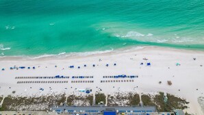 Vue aérienne d’une plage avec des rangées de parasols bleus et de chaises longues, un rivage sablonneux et une mer turquoise aux eaux limpides.
