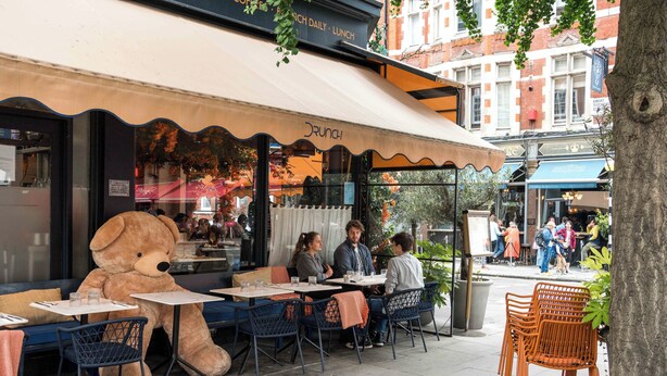 Un café en plein air avec des tables et des chaises, une grande décoration en forme d’ours en peluche et une enseigne sur laquelle on peut lire « CRUNCH ».