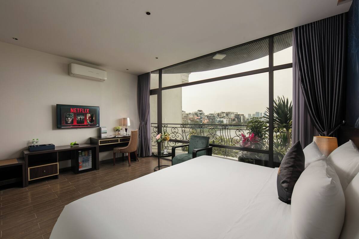 A hotel room with a large bed, a desk with a chair, a small table with a vase, and a TV on the wall.
