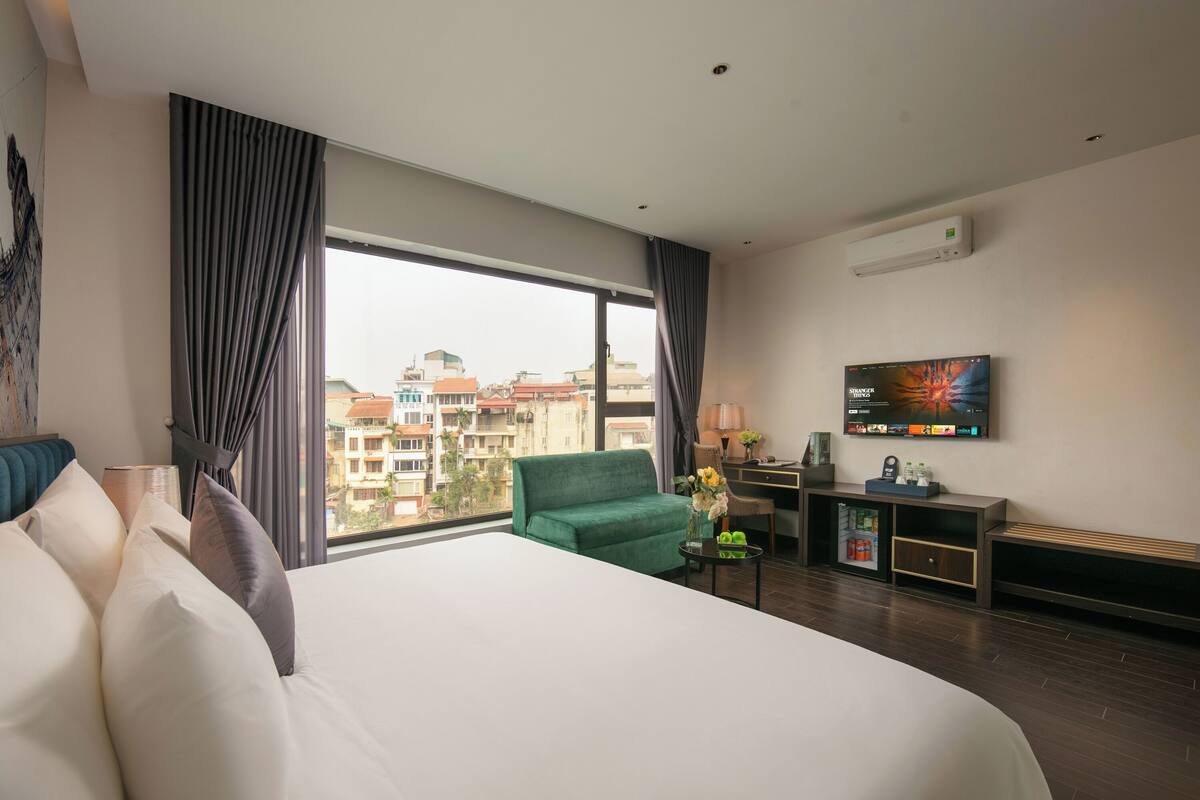 A hotel room with a large bed, a green armchair, a desk with a TV, and a view of buildings through the window.