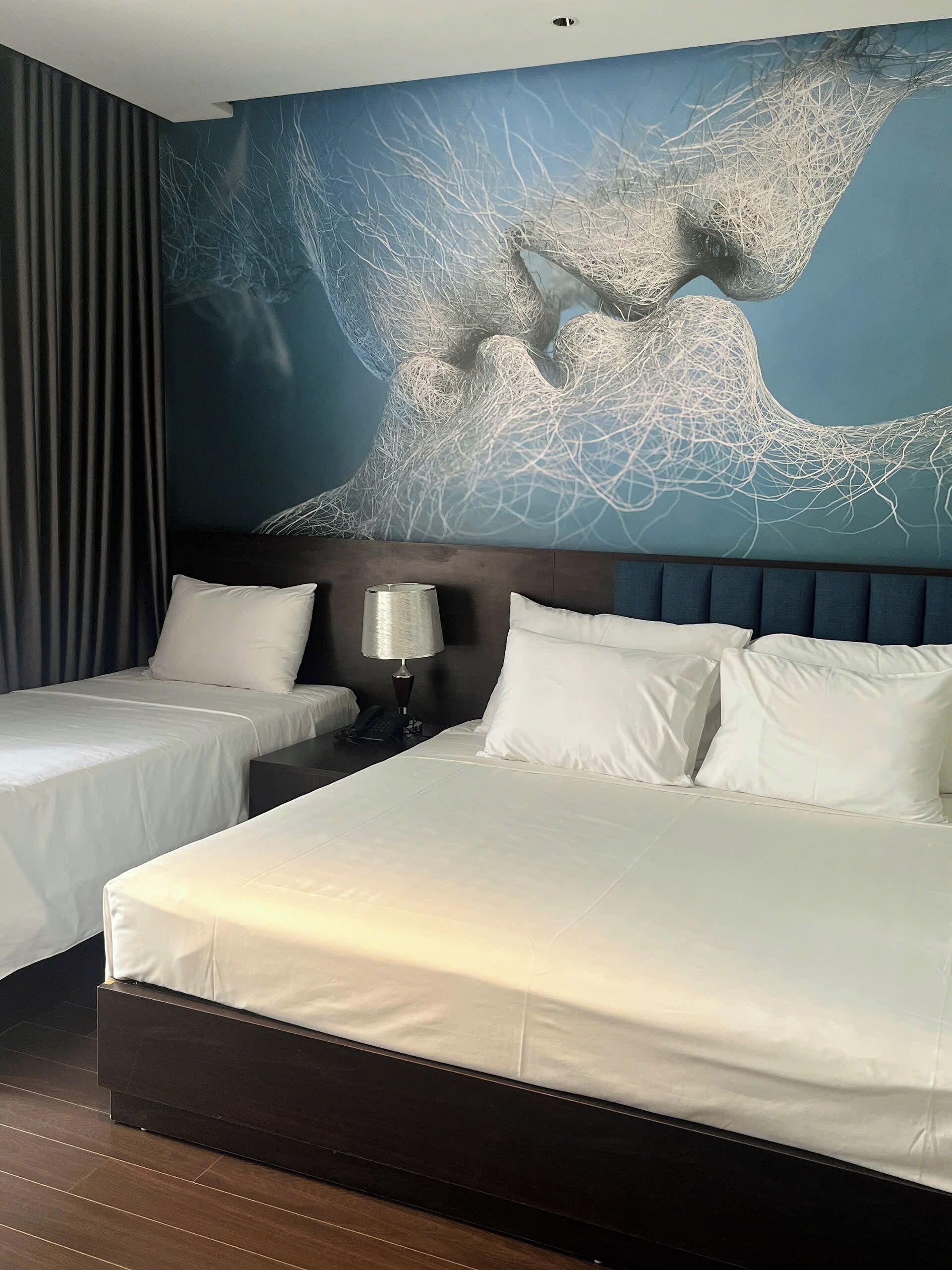 Une chambre d’hôtel avec deux lits, une grande fresque murale abstraite, une tête de lit sombre et une table de chevet avec une lampe.