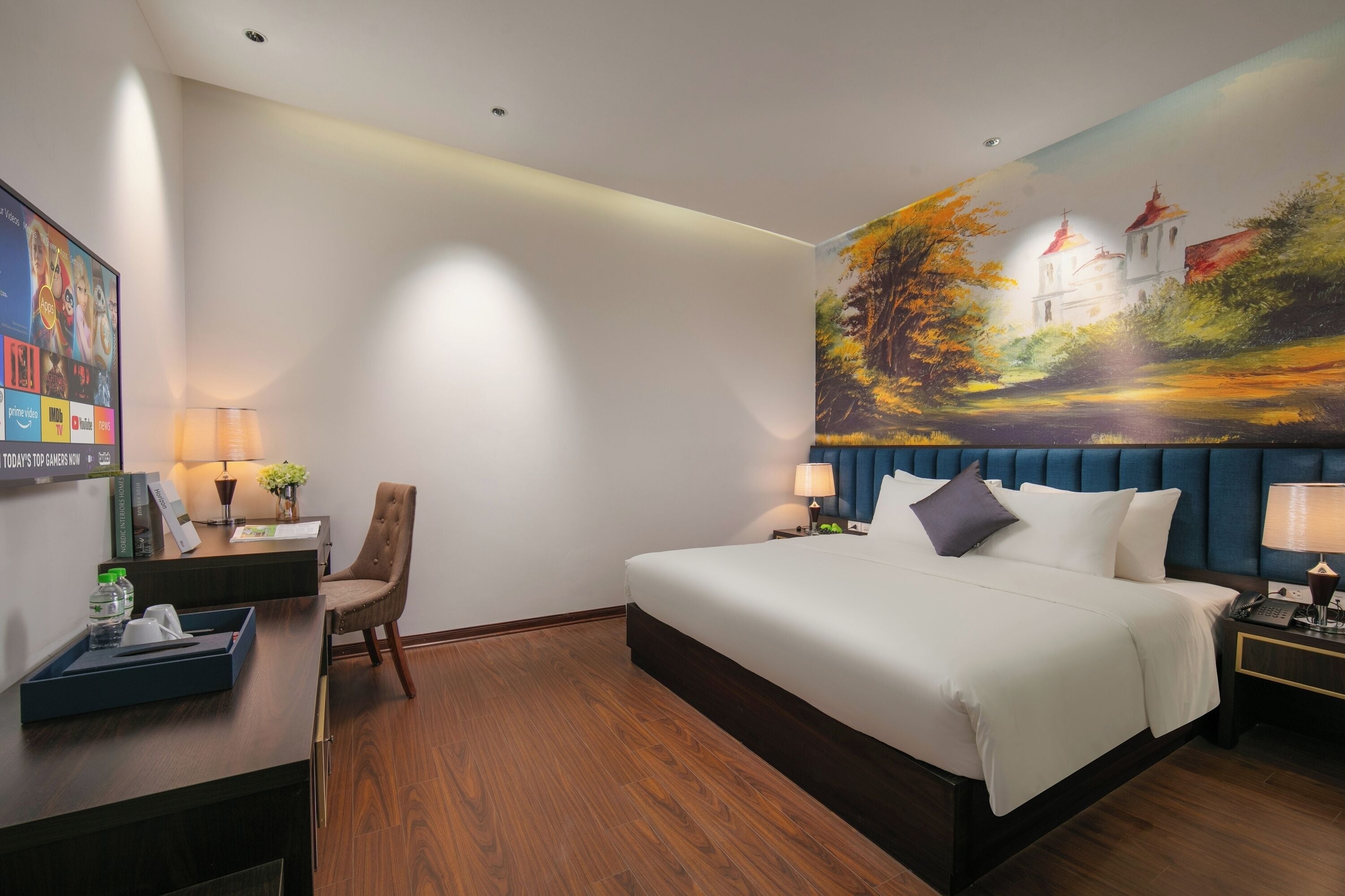 A hotel room with a large bed, a desk with a chair, a painting on the wall, and a TV on the wall.