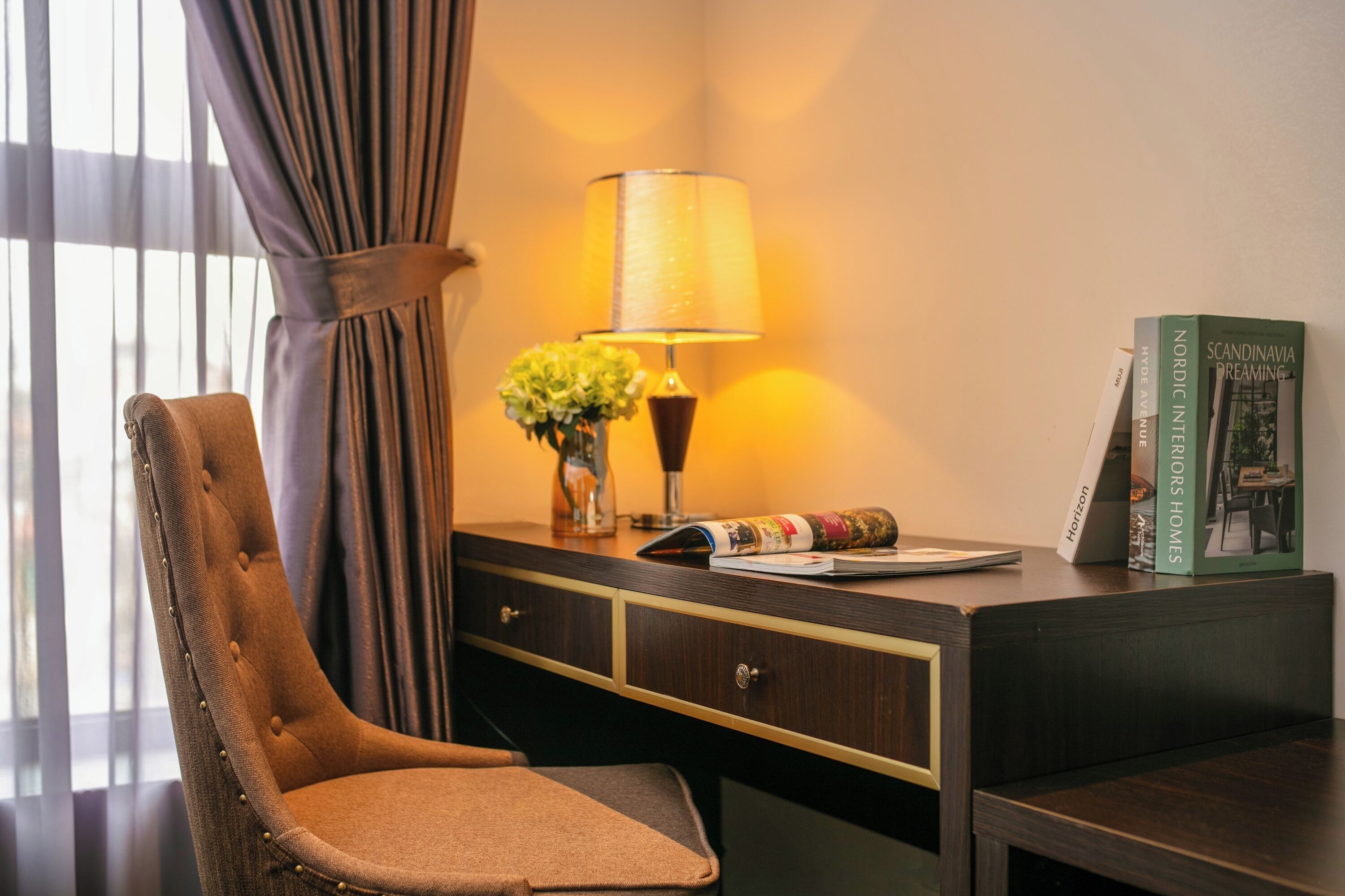 Une chambre d’hôtel avec un fauteuil à capitons, un bureau avec une lampe, un vase de fleurs et un livre intitulé « Intérieurs scandinaves ».