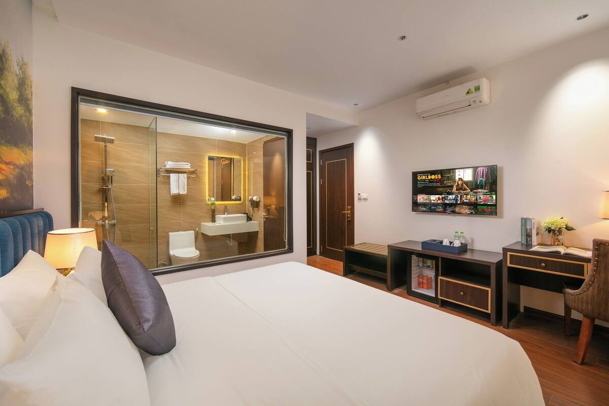 A hotel room with a large bed, a desk, a chair, a TV, a bathroom with a glass shower enclosure, and a wall-mounted air conditioner.
