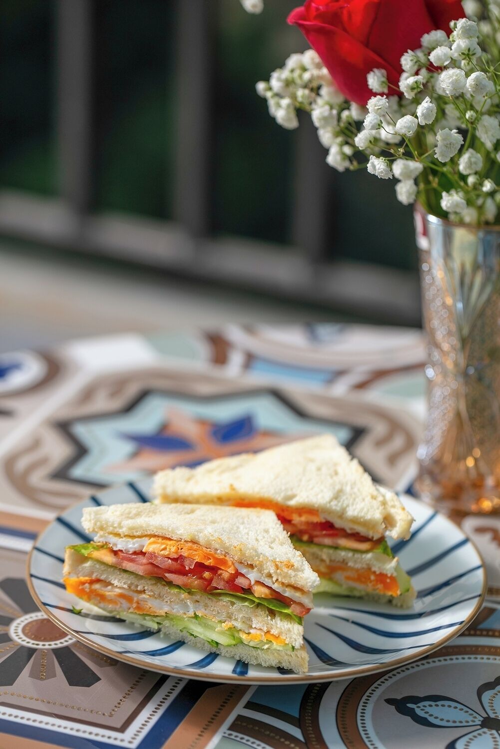 A sandwich on a plate with a decorative tablecloth, a vase with flowers, and a window in the background.