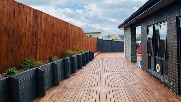 Una terraza de madera con macetas negras, un muro de ladrillo y cielo despejado.