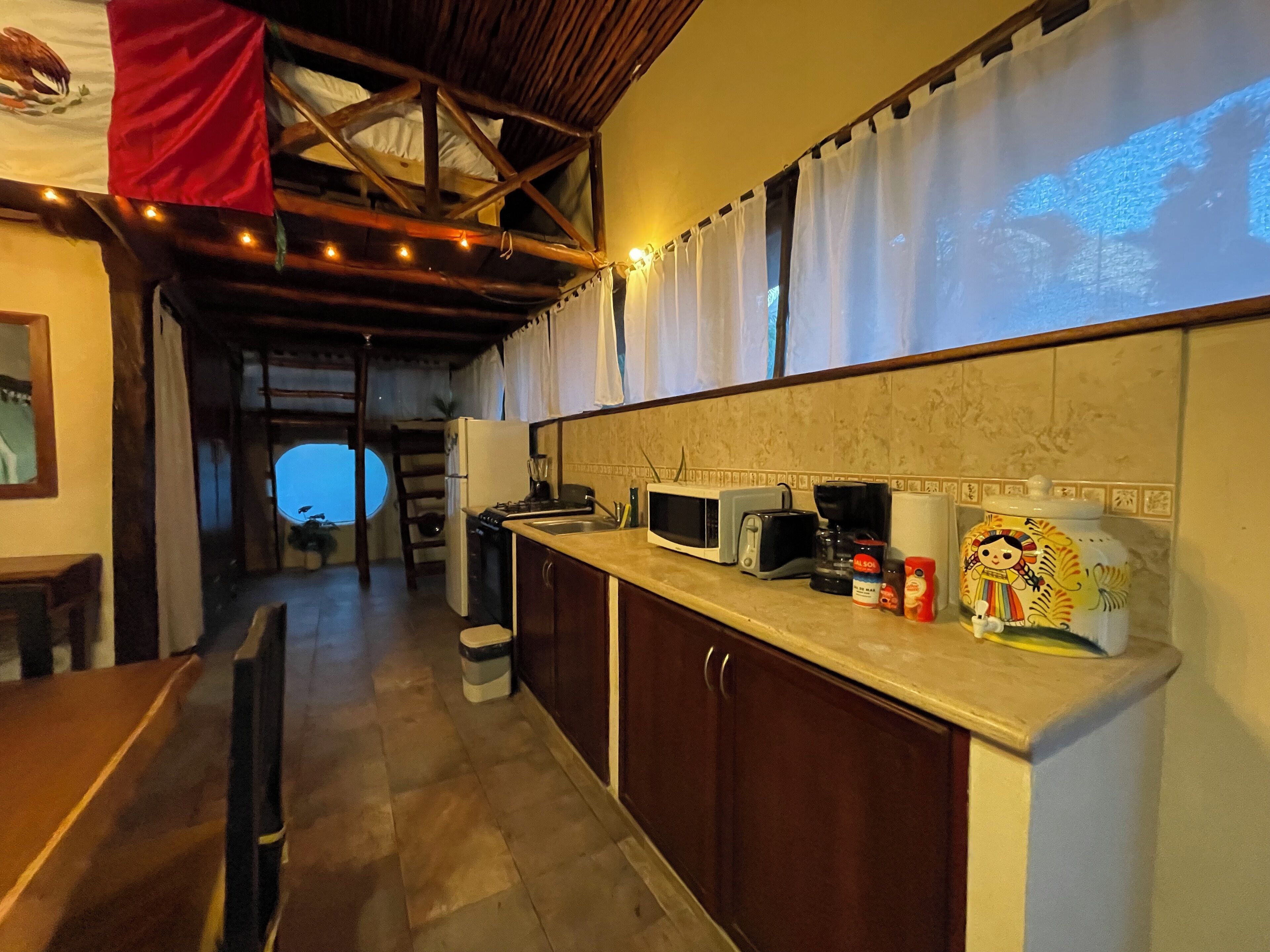 A kitchen with wooden cabinets, a microwave, toaster, and a coffee maker. There is a window with curtains and a view of the outside.