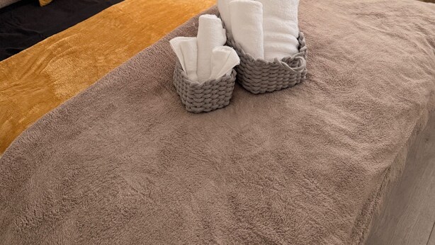 A neatly made bed with a brown blanket, two pillows, and two baskets containing folded towels. - Cozy one bed Apartment Near Heathrow (Slough)