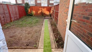 A residential backyard with a paved pathway, a small grassy area, and a brick building in the background. - Cozy one bed Apartment Near Heathrow (Slough)