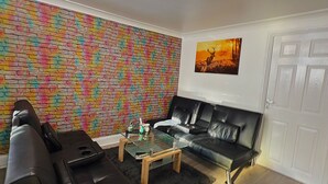 A living room with a black leather sofa, a glass coffee table, and a colorful brick-patterned wall. - Cozy one bed Apartment Near Heathrow (Slough)