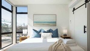 A bedroom with a large bed, a view of the beach through a window, and a painting on the wall. - Walk to the Beach & Pier! | Ocean View Roof Deck (Oceanside)