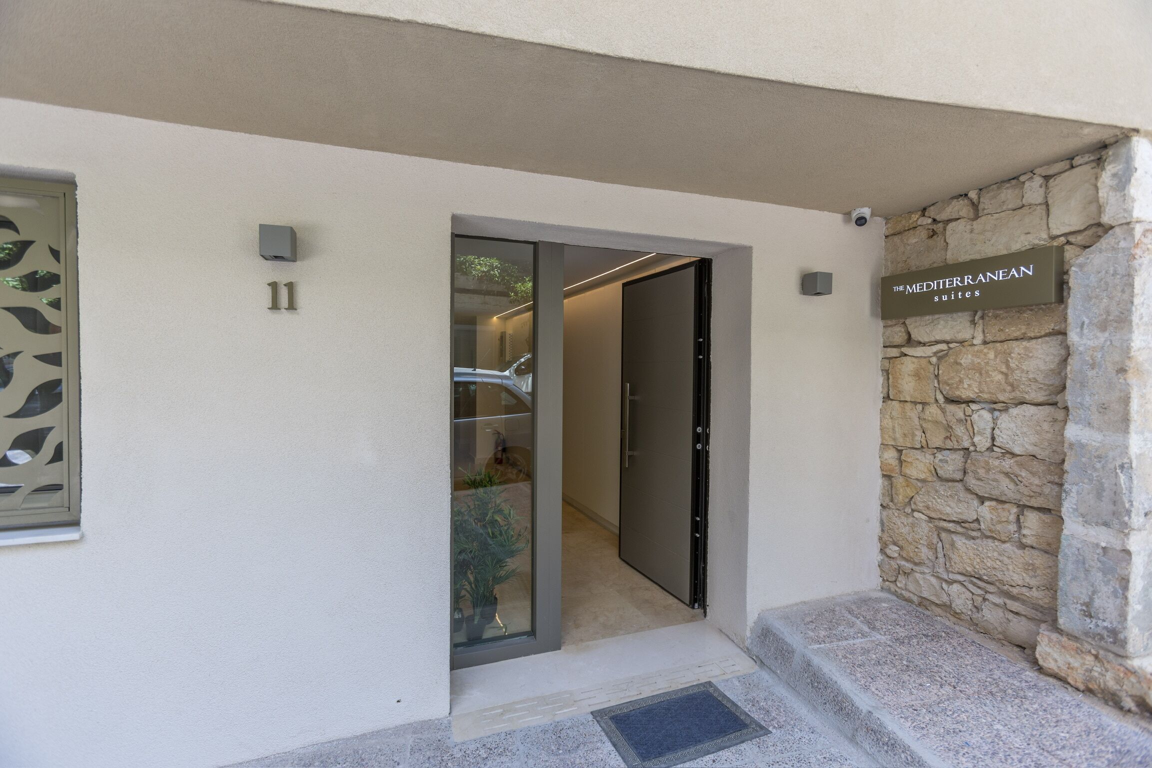 A modern building entrance with a stone wall, a glass door, and a sign for 'The Mediterranean Suites'.
