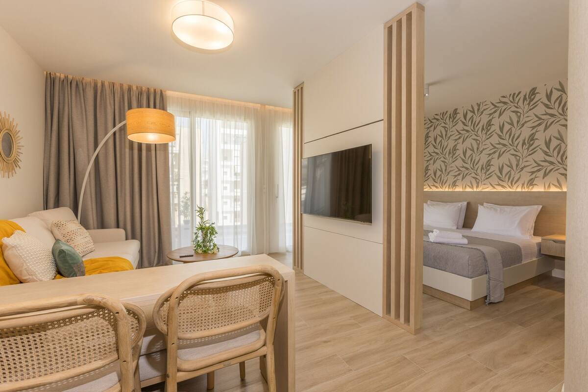 Una habitación de hotel moderna con zona de comedor, sofá, televisor y cama.