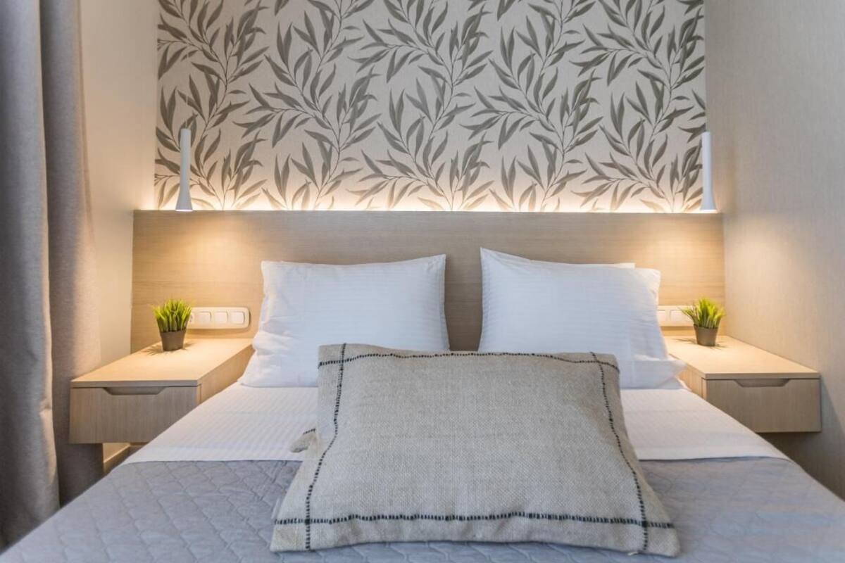Una cama bien hecha con sábanas blancas y una almohada decorativa, flanqueada por dos mesitas de noche con pequeñas plantas en macetas.