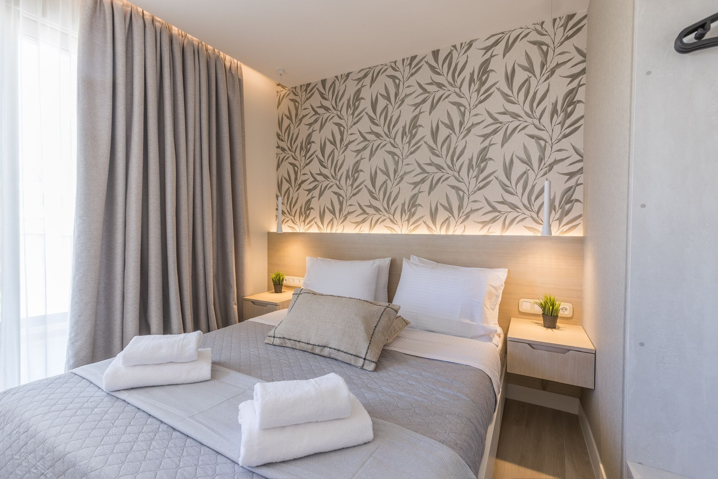 A neatly made bed with pillows and folded towels, a bedside table with a plant, and a wall with a leaf pattern.
