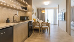 Una cocina moderna equipada con lavavajillas, microondas y fregadero. En la zona de comedor hay dos sillas de mimbre y una mesa pequeña. La habitación tiene un ventanal con cortinas y un televisor de pantalla plana montado en la pared.