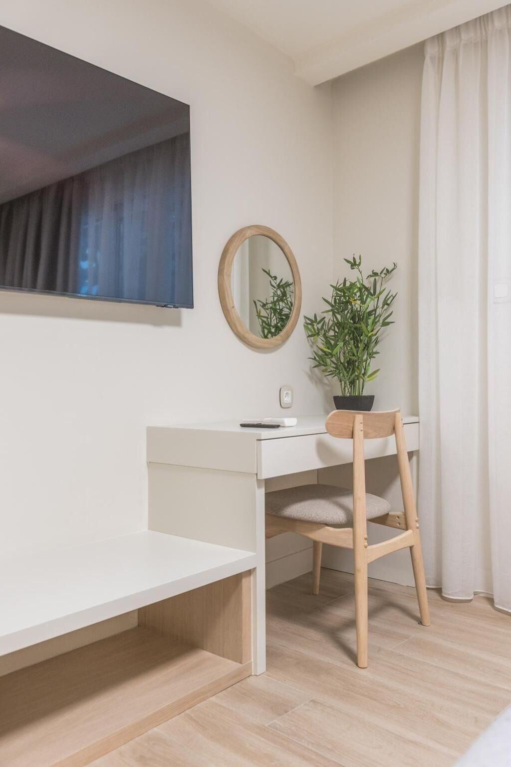 Ein schlicht eingerichtetes Zimmer mit weißem Schreibtisch, einem Holzstuhl, einem Spiegel und einem Blumentopf.