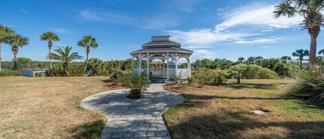 A gazebo with a white roof and open sides, surrounded by a paved pathway and palm trees.