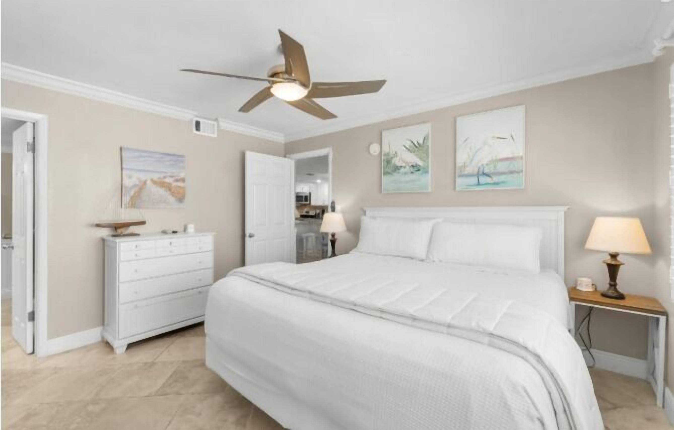 A bedroom with a large bed, a ceiling fan, a dresser, and two wall-mounted lamps.
