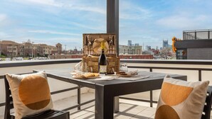 Terraza en la azotea con una mesa para dos, que incluye una botella de vino, copas y aperitivos. La terraza tiene vistas a un paisaje urbano con edificios y un cielo despejado.