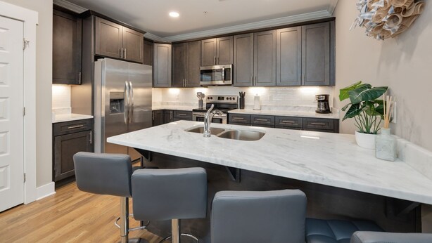 A modern kitchen with dark cabinets, a stainless steel refrigerator, a microwave, and a large island with a sink. There are bar stools and a potted plant on the island.