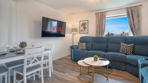 A modern living room with a blue sofa, a dining table with chairs, a TV mounted on the wall, and a small round table with a plant. - Golf Road Apartment | Grampian Lettings (Aberdeen)