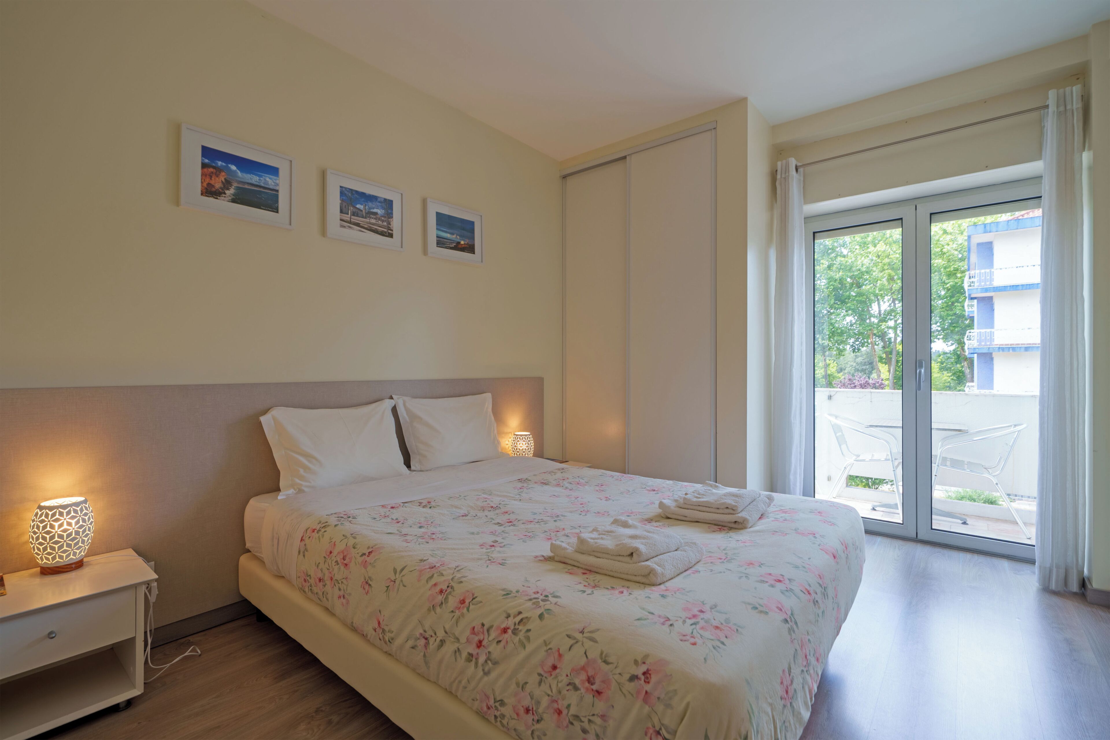 A hotel room with a bed, a nightstand, a lamp, and a view of the outside.