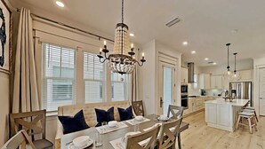 Un comedor amplio con una gran mesa de madera dispuesta para cuatro comensales, rodeada de sillas de madera. Un candelabro cuelga del techo y la habitación tiene ventanas grandes con persianas.