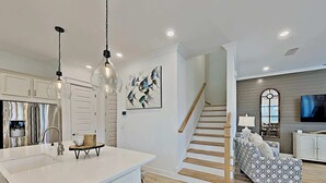 Una cocina moderna con armarios blancos, una isla grande e iluminación colgante. Una escalera conduce a una sala de estar con un sofá y un televisor.
