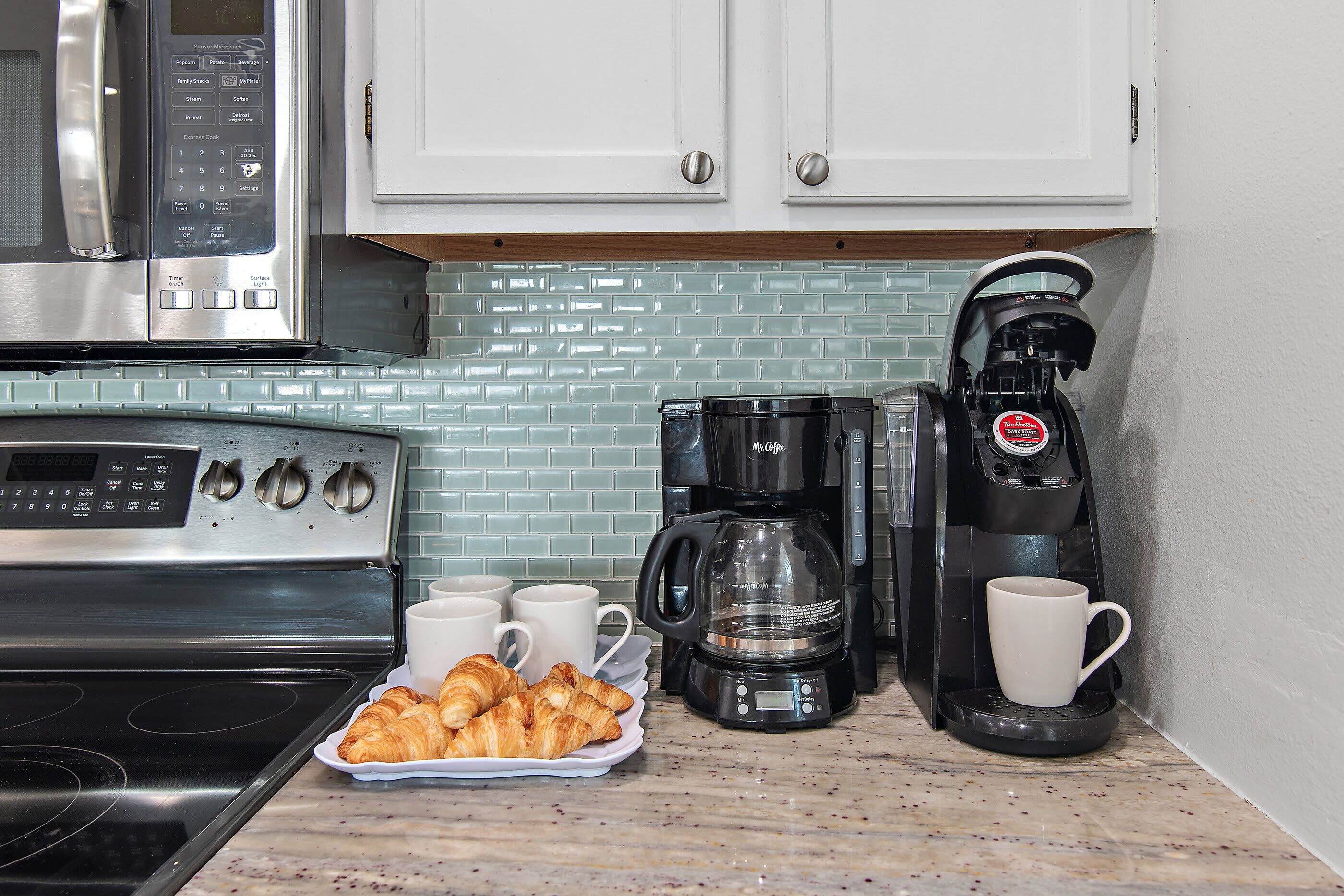 A kitchen with a stainless steel microwave, a coffee maker, and a croissant tray.