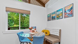 A dining area with a round glass table, blue chairs, a beige sofa with a white fur throw, and a window with a view of greenery.