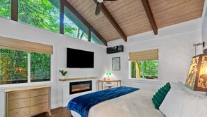 A bedroom with a large bed, a fireplace, a TV, and large windows with views of trees.
