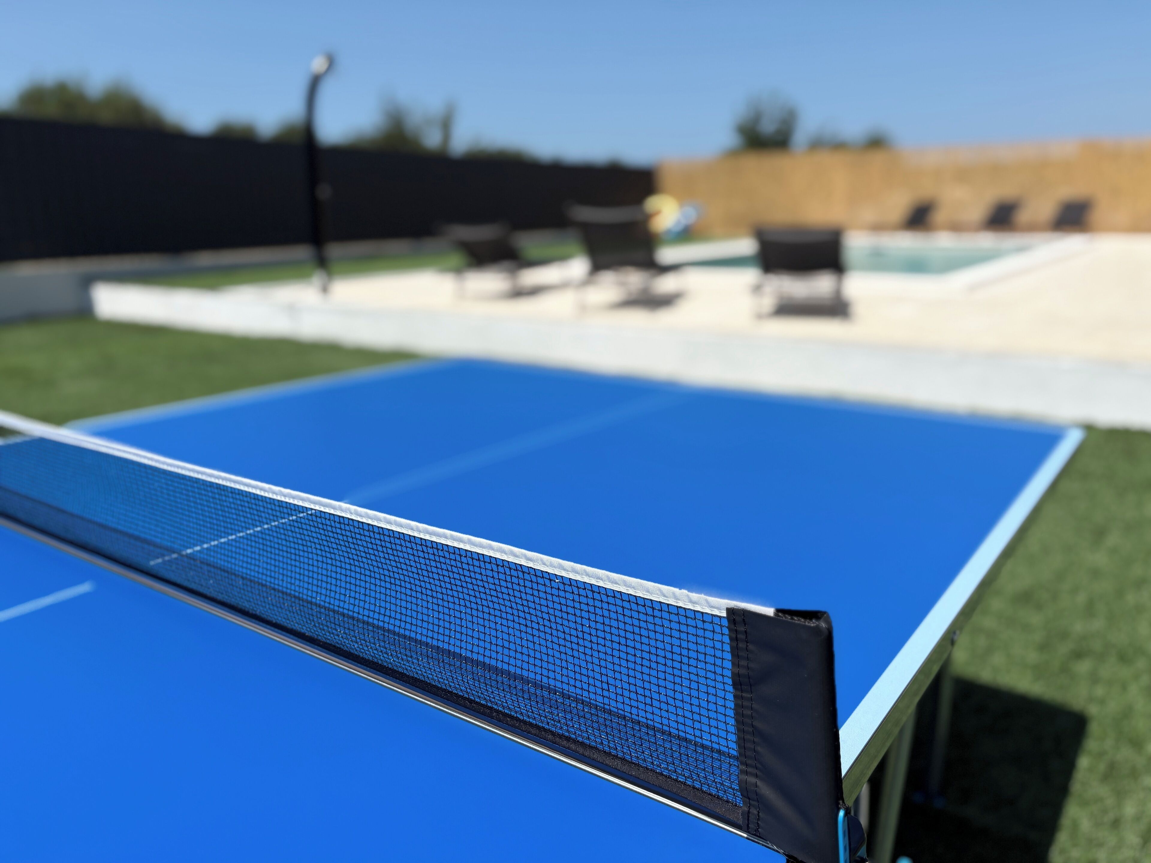 A blue ping pong table in an outdoor setting with a pool and lounge chairs in the background.