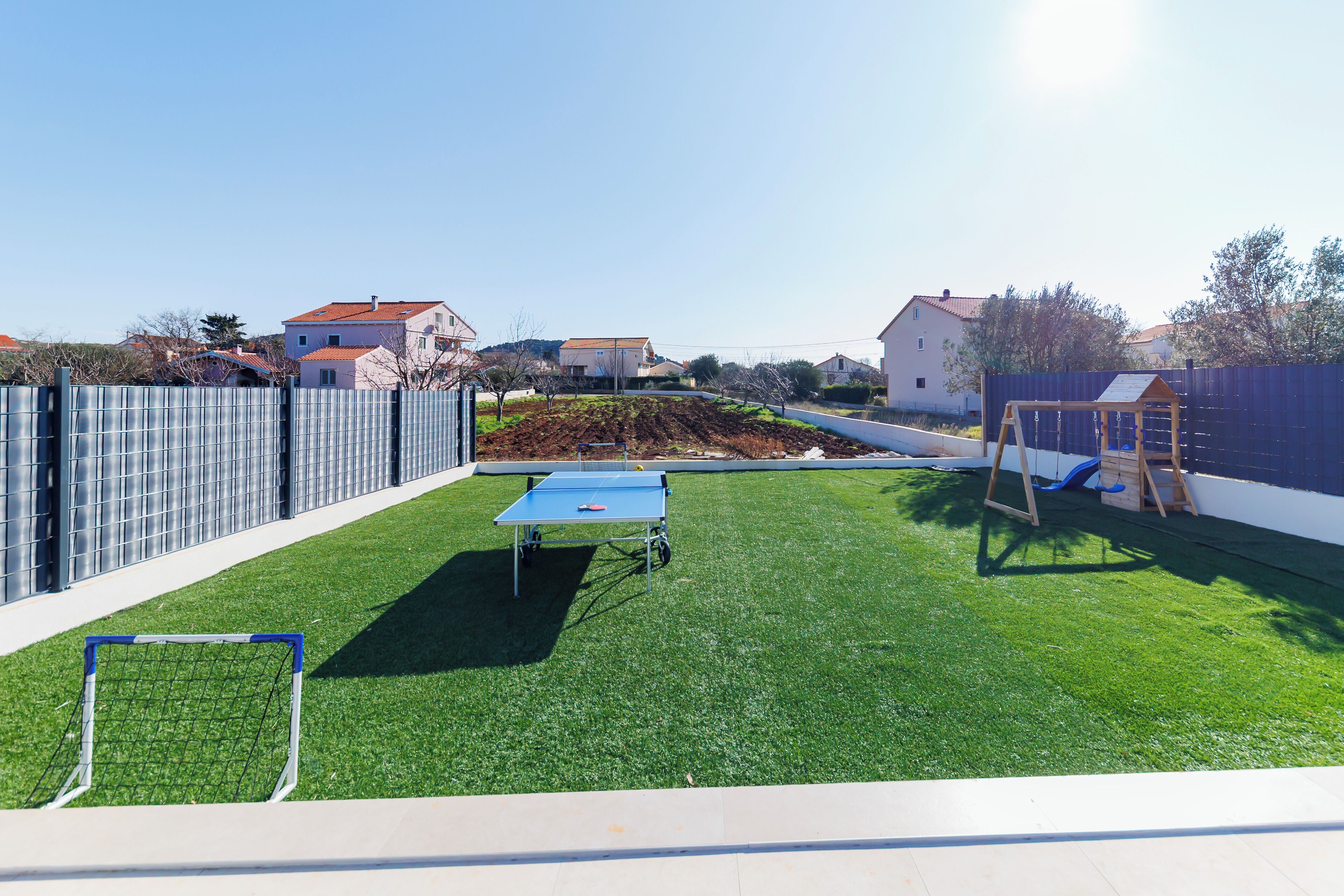 Un jardin avec une table de ping-pong bleue, un petit but de foot et une balançoire en bois.