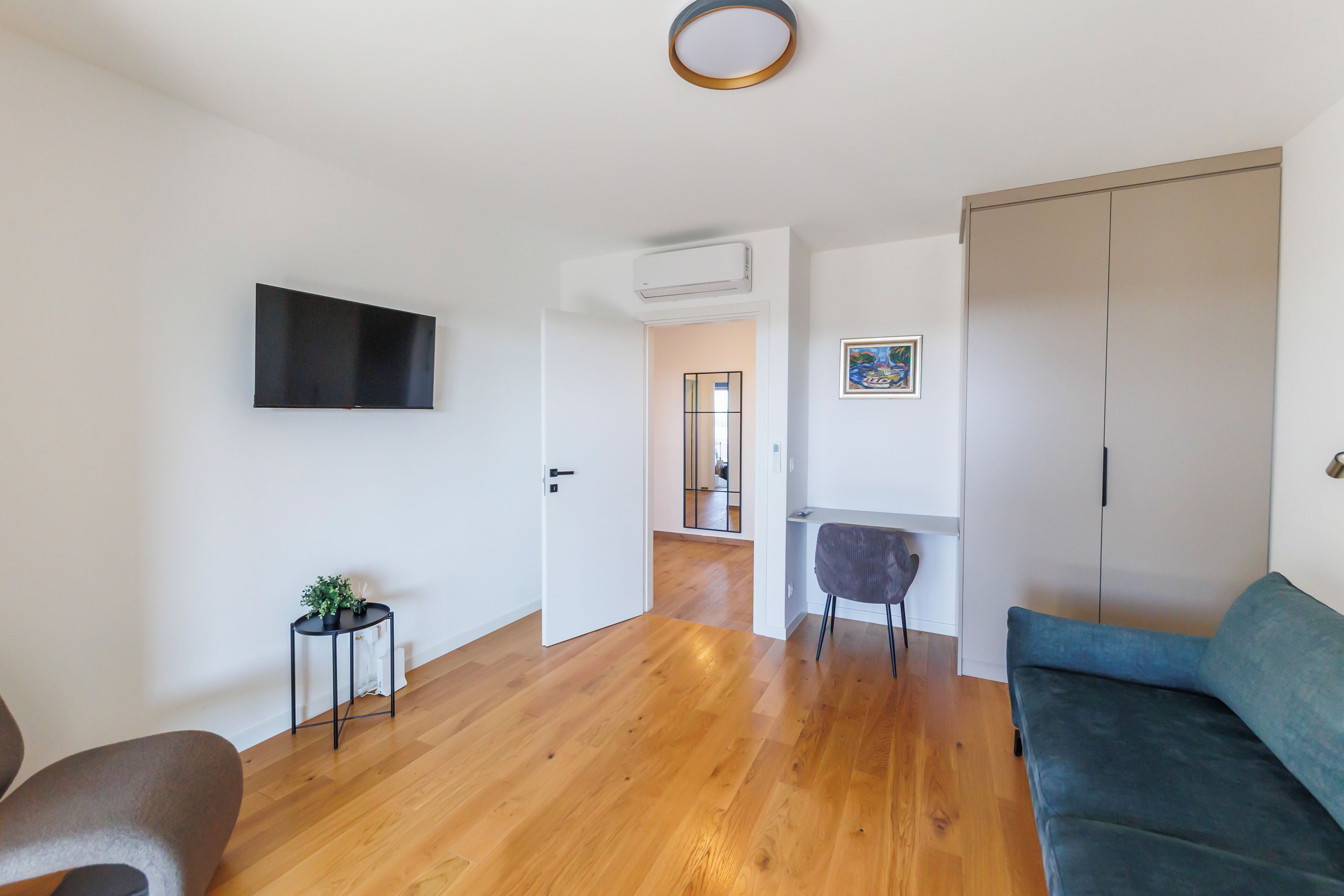 A modern living room with a wooden floor, a blue sofa, a small side table with a plant, a flat-screen TV mounted on the wall, and a wardrobe.
