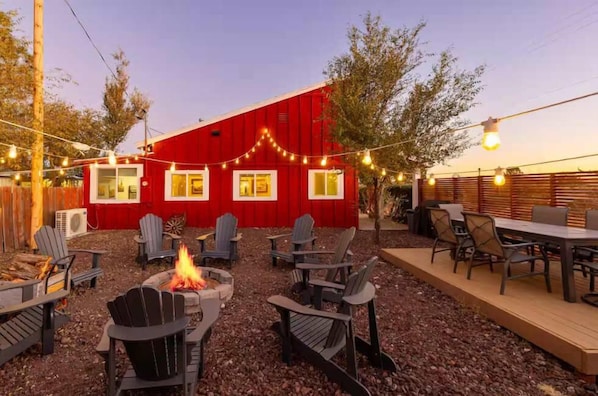 Un granero rojo con luces colgantes, mobiliario exterior con sillas Adirondack y una chimenea.