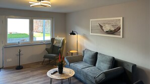 Een woonkamer met een grijze bank, een schommelstoel, een ronde salontafel en een lijst met een afbeelding aan de muur.