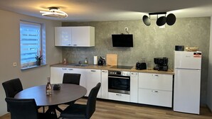 Een moderne keuken met witte kasten, een ingebouwde oven, een magnetron en een koelkast. Er is een eettafel met stoelen en een raam met jaloezieën.